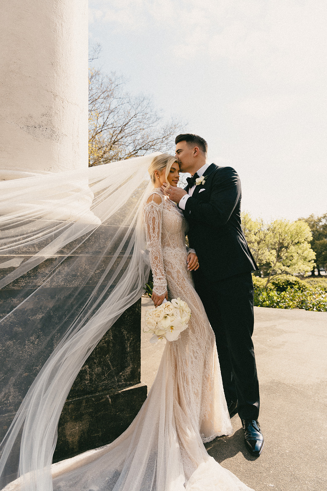 A bride and groom pose for their wedding portraits as her veil sways in the wind.