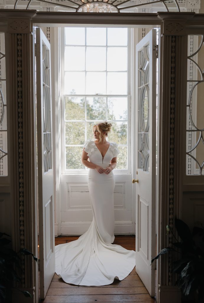 Bride in a fitted gown standing in a sunlit doorway at the Governor Thomas Bennett House, her train spread across the wood floor
